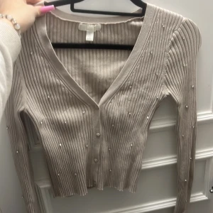 Beige ribbad kofta med diamanter  - Säljer en stilren beige ribbad kofta med pärldetaljer. Koftan har en v-ringad design och knappar framtill. Perfekt för att ge en elegant touch till din outfit.