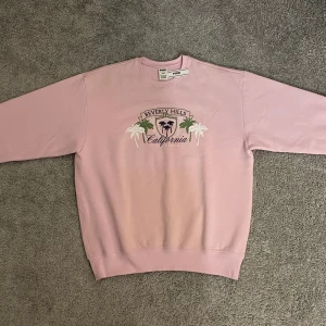 Rosa sweatshirt från PINK - Säljer en rosa sweatshirt från PINK med tryck av "Beverly Hills California" och palmer på framsidan. Tröjan har långa ärmar, en rund halsringning och är aldrig använd. Den är dessutom supermjuk inuti. Nypris 545 kr men säljer för 450