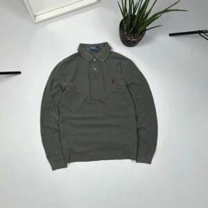 Grå pikétröja från Ralph Lauren - Snygg grå pikétröja från Ralph Lauren med klassisk krage och knappar. Den har långa ärmar och en liten röd logotyp på bröstet. Perfekt för en stilren look.