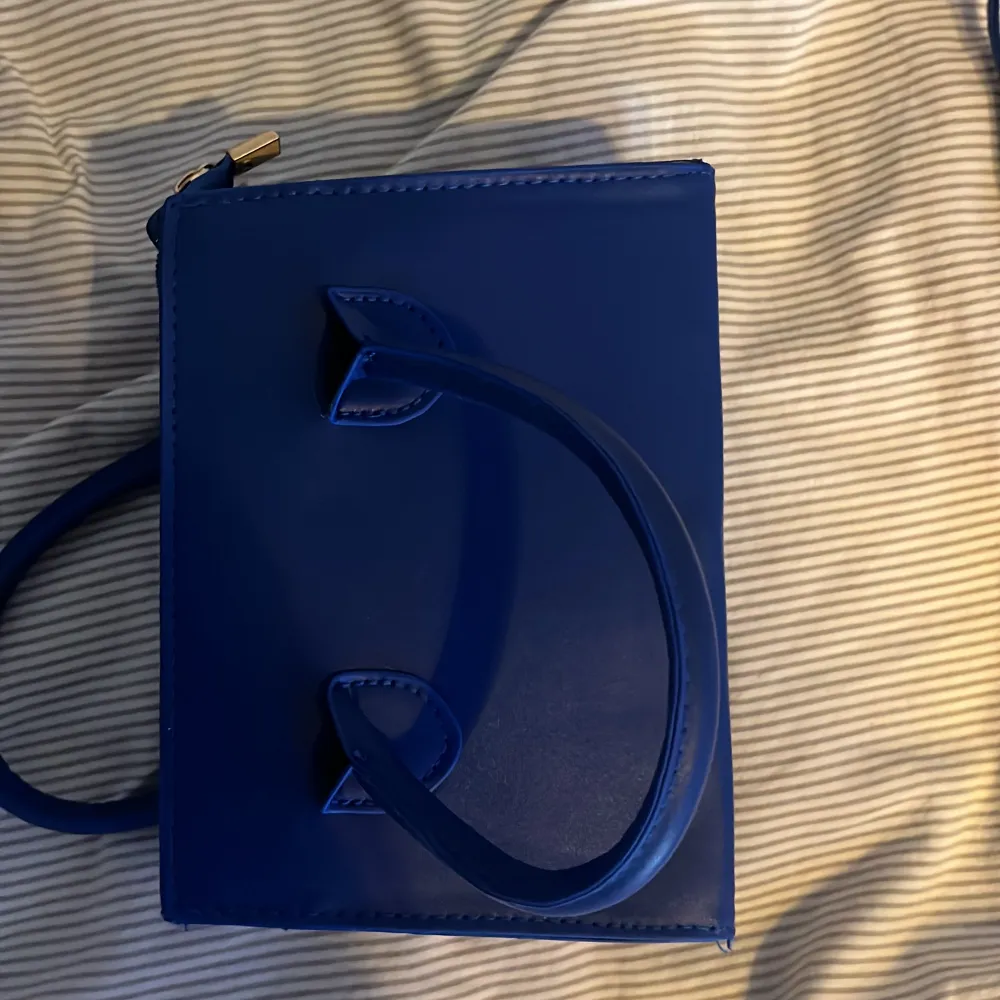 Snygg blå axelväska i syntetmaterial med både handtag och en lång axelrem. Väskan har en praktisk dragkedja upptill för säker förvaring. Perfekt för att bära med sig det viktigaste med stil.. Laukut & Käsilaukut.