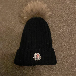 Svart ribbad mössa med pälsboll från Moncler - Snygg svart ribbad mössa från Moncler med en fluffig pälsboll på toppen. Perfekt för att hålla värmen under kyliga dagar. Den har en broderad logotyp framtill.