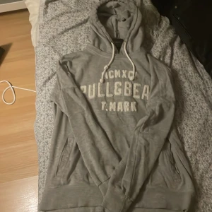 Grå hoodie från Pull&Bear Passar M - Säljer en grå hoodie från Pull&Bear med tryck på framsidan. Den har en klassisk design med justerbar huva och fickor framtill. Perfekt för en avslappnad stil.