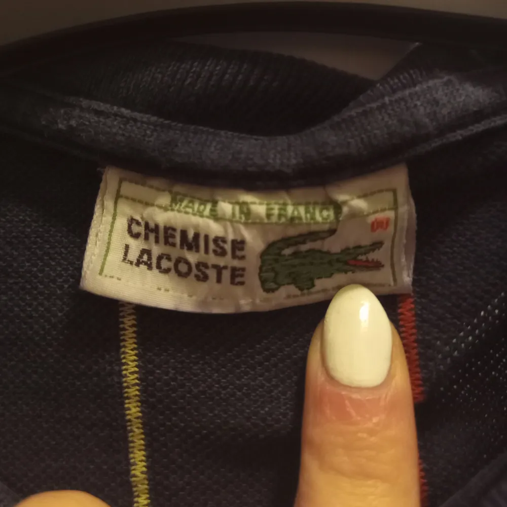 Snygg mörkblå pikétröja från Lacoste med diskreta ränder i olika färger. Tröjan har korta ärmar och klassisk krage med knappar. Perfekt för en stilren och avslappnad look.. Neuletakit & Villapaidat.