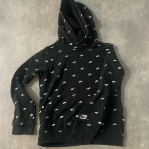 Svart hoodie med mönster från Nike - Säljer en svart hoodie från Nike med ett coolt mönster av små vita logotyper över hela tröjan. Perfekt för en avslappnad stil med en bekväm passform och huva.