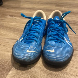 Blå Nike futsal skor  - Snygga blå Nike sneakers med vit logga och textdetaljer på hälen. Skorna har snörning och en sportig design, perfekt för en aktiv livsstil.