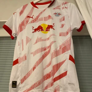  RB Leipzig fotbollströja från Puma - Snygg vit och röd RB Leipzig fotbollströja från Puma med korta ärmar. Tröjan har ett dynamiskt rött mönster och RB Leipzig-logga på bröstet. Perfekt för fans som vill visa sitt stöd med stil! 🏆