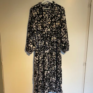 Blommig klänning från Lindex - Svart klänning från Lindex med ett blommigt mönster i vitt och gult. Klänningen har långa ärmar och ett knytband i midjan samt knappar i fram.