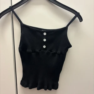 Svart ribbad linnetopp med knappar - Snygg svart ribbad linnetopp med smala axelband och tre dekorativa knappar framtill. Det står att stl är 12-13 år men tycker den passar mindre barn också.