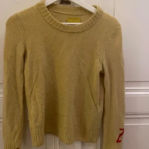 Beige tröja från Zadig & Voltaire - Snygg beige tröja från Zadig & Voltaire med ribbade detaljer vid hals och ärmslut. Den har en diskret röd detalj på ärmen som ger en cool touch. Perfekt för en stilren look.