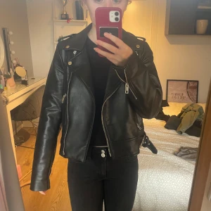 Svart skinnjacka med dragkedjor - Snygg svart skinnjacka med coola dragkedjor och knappar. Jackan har en klassisk bikerstil med bälte i midjan och långa ärmar. Perfekt för en edgy look!