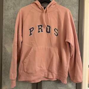 Rosa hoodie från Canard - Säljer en rosa hoodie från Canard med texten 'PROS' i svart på bröstet. Den har en klassisk känguruficka och justerbar huva. Perfekt för en avslappnad stil.