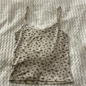 Blommig linnetopp från Brandy Melville - Söt linnetopp från Brandy Melville i beige med små blommor i blått. Toppen har spetsdetaljer vid halsringningen och en liten rosett framtill. Perfekt för en avslappnad stil.