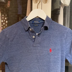 Blå pikétröja från Ralph Lauren - Snygg blå pikétröja från Ralph Lauren med klassisk krage och två knappar. Den har en röd broderad logotyp på bröstet och är i en slim passform. Perfekt för en stilren look!