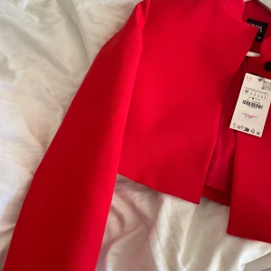 Röd kavaj från Zara - Snygg röd kavaj från Zara med en modern, croppad design. Den har en stilren look med svarta knappar och en enkel krage. Perfekt för att ge en färgklick till din outfit.