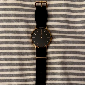 Elegant klocka med svart läderarmband - Klocka från Daniel Wellington med svart urtavla och guldfärgad boett. Behöver nytt batteri. Priset går att diskutera.