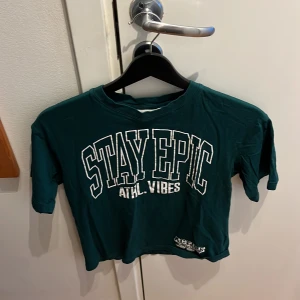 Grön t-shirt med tryck från H&M - Cool grön t-shirt från H&M med texten 'STAY EPIC ATHL. VIBES' i vitt tryck på framsidan. Perfekt för en avslappnad stil. Kortärmad och i mjukt material.