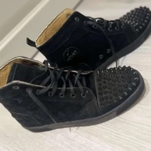 Svarta sneakers med nitar - Snygga svarta sneakers med nitar på tån och snörning. Skorna har en röd sula och är tillverkade i mocka. Perfekta för att ge en edgy touch till din stil.