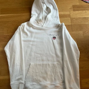 Vit hoodie från GANT - Säljer en stilren vit hoodie från GANT med broderad logga och emblem på bröstet. Tröjan har en klassisk känguruficka och ribbade muddar vid ärmslut och nederkant. Perfekt för en avslappnad look.