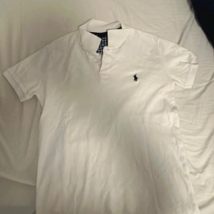Vit pikétröja från Ralph Lauren - Säljer en klassisk vit pikétröja från Ralph Lauren med korta ärmar och den ikoniska loggan broderad på bröstet. Kragen har ett stilrent rutigt mönster i blått och vitt. Perfekt för en avslappnad men stilren look.