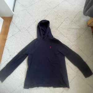 Mörkblå hoodie från Polo Ralph Lauren - Säljer en stilren mörkblå hoodie från Polo Ralph Lauren med det klassiska röda logotypen på bröstet. Tröjan har långa ärmar och en bekväm huva. Perfekt för en avslappnad stil. Original pris 1166kr
