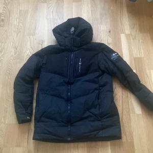 Sail racing jacka  - Säljer en svart dunjacka från sail racing med huva och dragkedja. Jackan har en broderad logga på bröstet och Perfekt för kalla dagar.