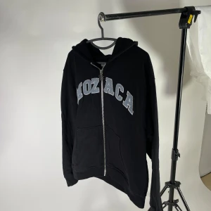 Svart hoodie från Mozaca  - Premiumhoodie från Mozaca kvitto och original förpackning tillkommer extremt skön och känns exklusiv. Säljer en svart hoodie från Mozaca med dragkedja och stor text i ljusblått på framsidan. Den har en cool detalj med en blomformad dragkedja och praktiska fickor framtill. Perfekt för en avslappnad stil. Hoodien är i nyskick