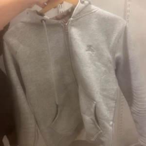 Grå hoodie från Burberry - Säljer en stilren grå hoodie från Burberry med dragkedja och justerbar huva. Tröjan har ett diskret broderat emblem på bröstet och klassiskt Burberry-mönster på insidan av huvan. Perfekt för en avslappnad och trendig look.