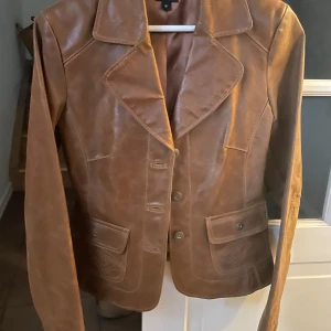 Brun skinnjacka med knappar - Snygg brun skinnjacka med klassisk krage och knappar framtill. Jackan har långa ärmar och två fickor med lock. Perfekt för en stilren look.