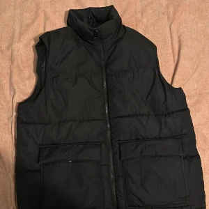 Svart dunväst med dragkedja - Säljer en svart dunväst med dragkedja och stora fickor framtill. Perfekt för kyligare dagar och enkel att matcha med andra plagg. Västen har en hög krage för extra värme. Obs att under höger fickan så finns det en liten skada men man märker knappt 