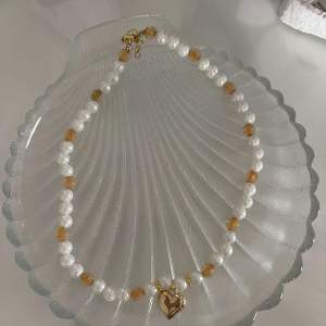 Ett elegant halsband med vita pärlor och gyllene detaljer. Det har små guldpärlor och ett hängande guldhjärta i mitten. Perfekt för att ge en touch av glamour till din outfit. Passar både till vardag och fest!