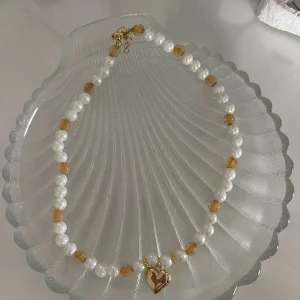 Halsband med vita pärlor och guldhjärta - Ett elegant halsband med vita pärlor och gyllene detaljer. Det har små guldpärlor och ett hängande guldhjärta i mitten. Perfekt för att ge en touch av glamour till din outfit. Passar både till vardag och fest!