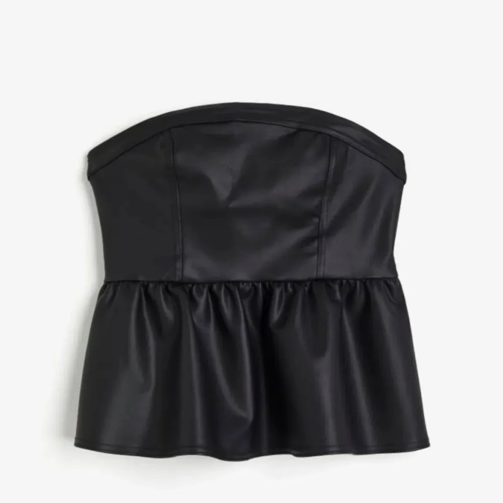 Snygg svart axelbandslös topp i skinnimitation med volangdetalj nedtill. Perfekt för en utekväll eller fest. Toppen har en tight passform upptill och en lösare volang som ger en fin siluett.. Yläosat.
