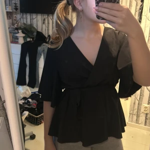 Svart blus med knytband - Säljer en elegant svart blus med knytband i midjan, aldrig använd. Blusen har korta ärmar och en v-ringad design som ger en stilren look. Perfekt för både vardag och fest!