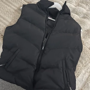 Svart dunväst från Zoul - Säljer en svart dunväst från Zoul i storlek M. Västen har en dragkedja framtill och är perfekt för kyliga dagar. Den är i mycket bra skick och passar bra till både höst och vinter. En stilren och praktisk väst för alla tillfällen.