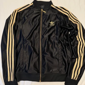 Svart Adidas jacka med guldiga ränder - Säljer en snygg svart Adidas jacka med guldiga ränder längs ärmarna. Den har en dragkedja fram och det klassiska Adidas-loggan på bröstet. Perfekt för både träning och vardag. Jackan är i mycket bra skick och passar perfekt till höst och vår. Ps liten i storlek! 