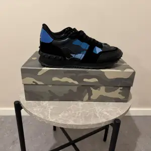 Säljer ett par snygga Valentino Rockrunner sneakers i svart och blått. Skorna har ett coolt camouflage-mönster och är i mycket bra skick. Perfekta för både vardag och fest! De har en bekväm passform med snörning och är gjorda i skinn och syntetmaterial.