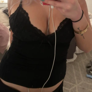 Svart spetslinne - Säljer ett snyggt svart linne med spetsdetaljer och smala axelband. Perfekt för en utekväll eller fest. Linnet har en liten rosett framtill och är i ett mjukt material som sitter skönt mot huden.