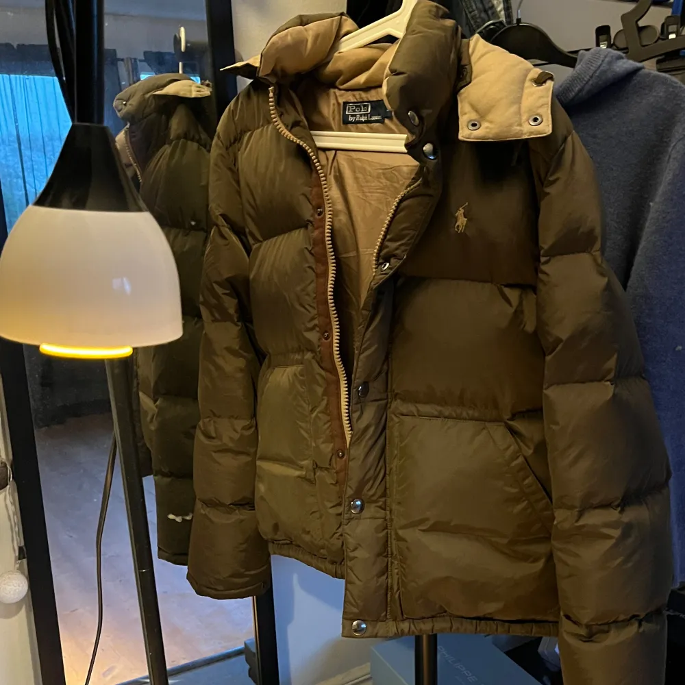 Säljer en snygg brun dunjacka från Ralph Lauren i mycket bra skick. Jackan har en klassisk design med dragkedja och knappar framtill. Perfekt för kalla höst- och vinterdagar. Den har långa ärmar och en mysig krage för extra värme. Passar perfekt för både vardag och lite finare tillfällen.. Jackor.