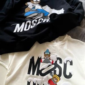 Svart och vit tröja från Moschino med nallemotiv - Två snygga tröjor från Moschino, en svart hoodie och en vit T-shirt, båda med coola nallemotiv. Den svarta har en nalle på en snöskoter och den vita har nallar med snödetaljer!🐻❄️T-shirten är aldrig använd= nyskick, hoodien är använd ett par gånger✨Nypris hoodie ca 1500:- Tshirt 599:-  Bud mottages men rimligt ca 50% på nypriset. Storlek 12 år. Paketpris 900:-