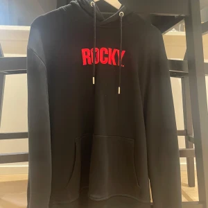 Svart hoodie med röd text - Säljer en svart hoodie med texten 'ROCKY' i rött på bröstet. Den har en klassisk design med dragsko i huvan och en stor ficka framtill. Perfekt för en avslappnad stil eller träning. Passar bra till både höst och vinter.