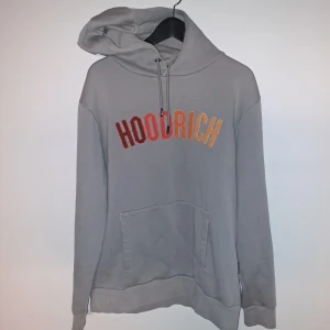 Grå hoodie från Hoodrich - Säljer en snygg grå hoodie från Hoodrich i storlek M. Den har en stor ficka fram och texten 'HOODRICH' i rött och orange på bröstet. Perfekt för en avslappnad stil eller att mysa i hemma. 🧡 betalning sker med swish