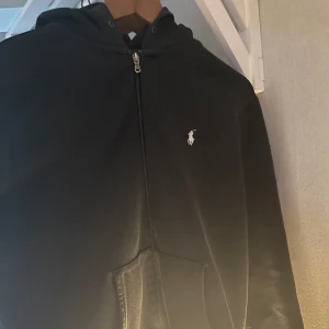 Svart hoodie från Ralph Lauren - Säljer en snygg svart hoodie från Ralph Lauren i mycket bra skick. Den har en dragkedja framtill och en klassisk logga på bröstet. Perfekt för höst och vår, med en skön passform och praktiska fickor. Perfekt för vardagsbruk eller en avslappnad stil.