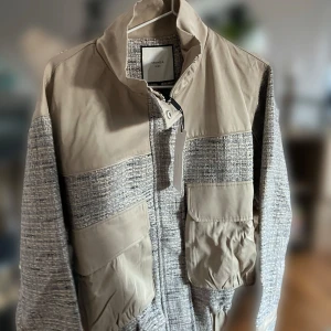 Beige och grå jacka från Neo Noir - Säljer en snygg jacka från Neo Noir i beige och grått. Jackan har en cool mix av material med stickade detaljer och släta partier. Den har en dragkedja framtill och praktiska fickor. Perfekt för höst och vår! Nypris 1499. Aldrig använd med prislapp kvar.