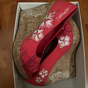 Rosa klackar från Ed Hardy - Säljer ett par unika rosa kilklackar från Ed Hardy med blommigt mönster. Skorna har en korkliknande sula och är dekorerade med vita och rosa blommor samt snygga detaljer på klacken. Perfekta för sommarens fester eller en dag när du vill sticka ut lite extra! 🌸