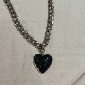 Silverfärgat halsband med svart hjärthänge - Snyggt och trendigt halsband i silverfärg med ett stort svart hjärthänge. Kedjan är grov och ger en cool look, perfekt för att piffa upp vilken outfit som helst. Passar både till vardags och fest. Ett måste för alla som älskar statement-smycken!