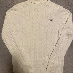 Vit kabelstickad tröja från Gant - Säljer en superfin vit kabelstickad tröja från Gant. Den har en klassisk polokrage och är perfekt för kyliga dagar. Tröjan är i mycket bra skick och har Gant-loggan broderad på bröstet. Perfekt för både höst och vinter!