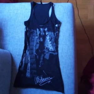 Svart linne med tryck från Madonna - Snyggt svart linne med ett coolt tryck av en person på framsidan. Perfekt för en avslappnad stil eller en utekväll. Linnet är ärmlöst och har en bekväm passform. Märket Madonna är tryckt på insidan.