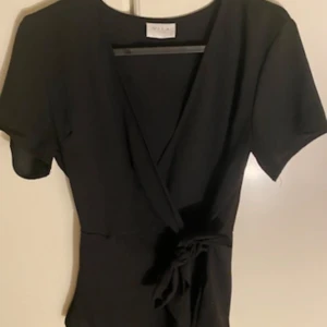 Svart omlottröja från Vila - Snygg svart omlottröja från Vila. Den har korta ärmar och ett knytband i midjan för en smickrande passform. Perfekt för både vardag och fest! 😍
