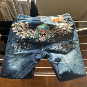 Kingz jeans med dödskallebroderi - Säljer ett par unika blå jeans med coolt dödskallebroderi och vingar på baksidan. Jeansen har knappar framtill och slitna detaljer för en edgy y2k look!