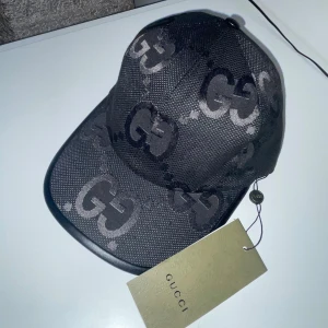 Svart keps från Gucci - Snygg svart keps från Gucci med det ikoniska GG-mönstret. Kepsen är i nytt skick och har en justerbar rem baktill för perfekt passform. Perfekt för att ge din outfit en lyxig touch!